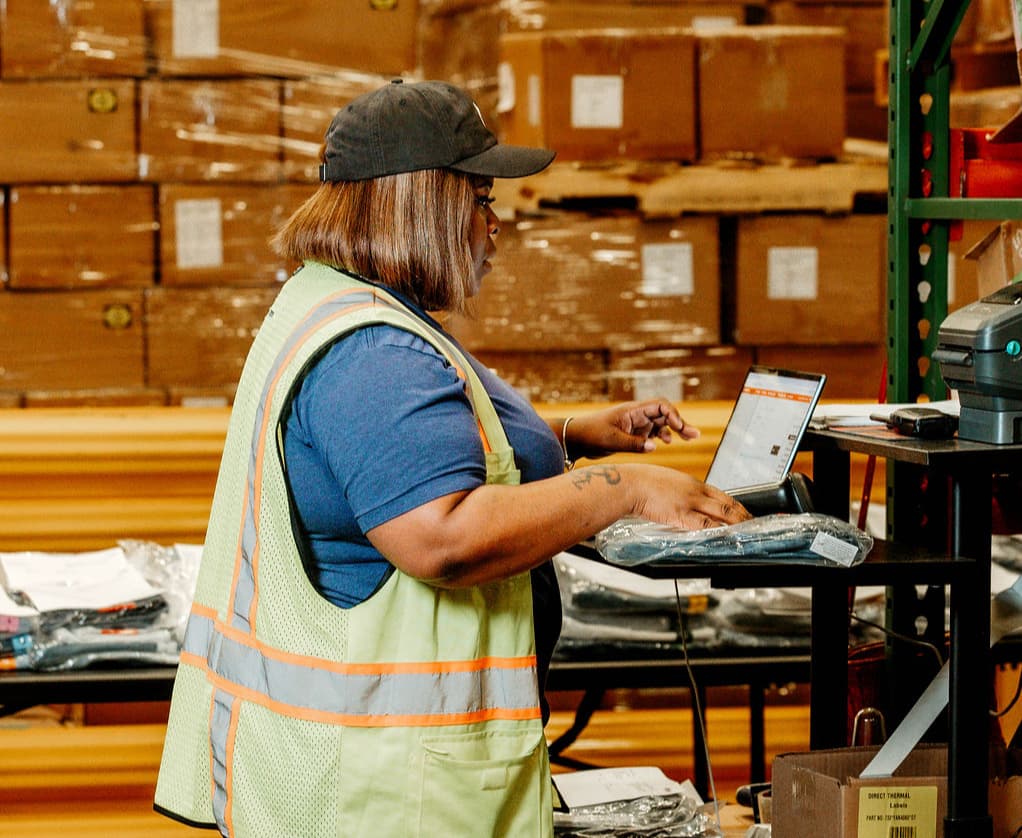 A warehouse worker in a reflective safety vest and cap stands at a workstation typing on a laptop with packaged items on the table and palletized boxes stacked behind them