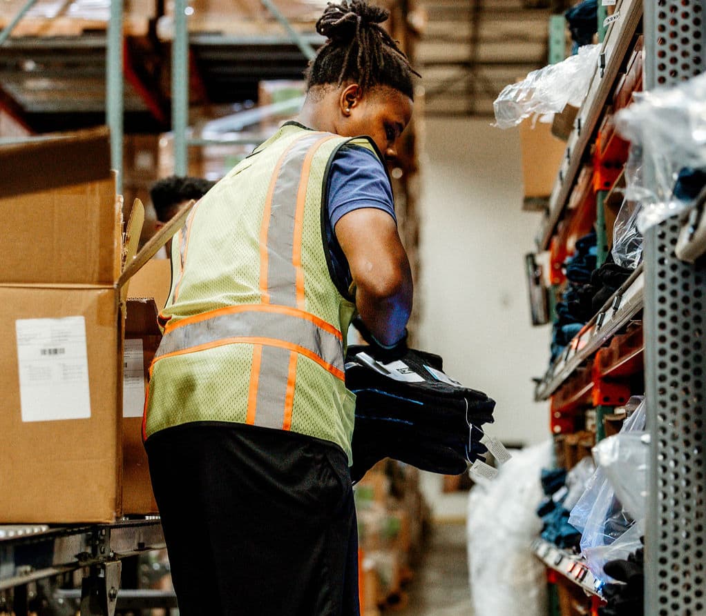 A warehouse worker in a reflective safety vest picks folded clothing items from a metal shelf in a busy warehouse aisle