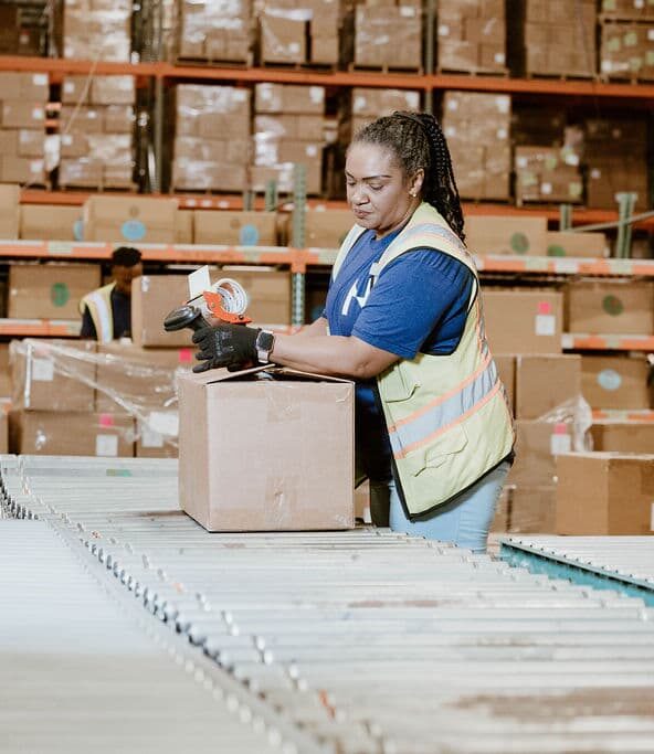 A warehouse worker in a reflective safety vest seals a cardboard box with a hand‑held tape dispenser on a roller conveyor amid rows of stacked boxes and pallet racks