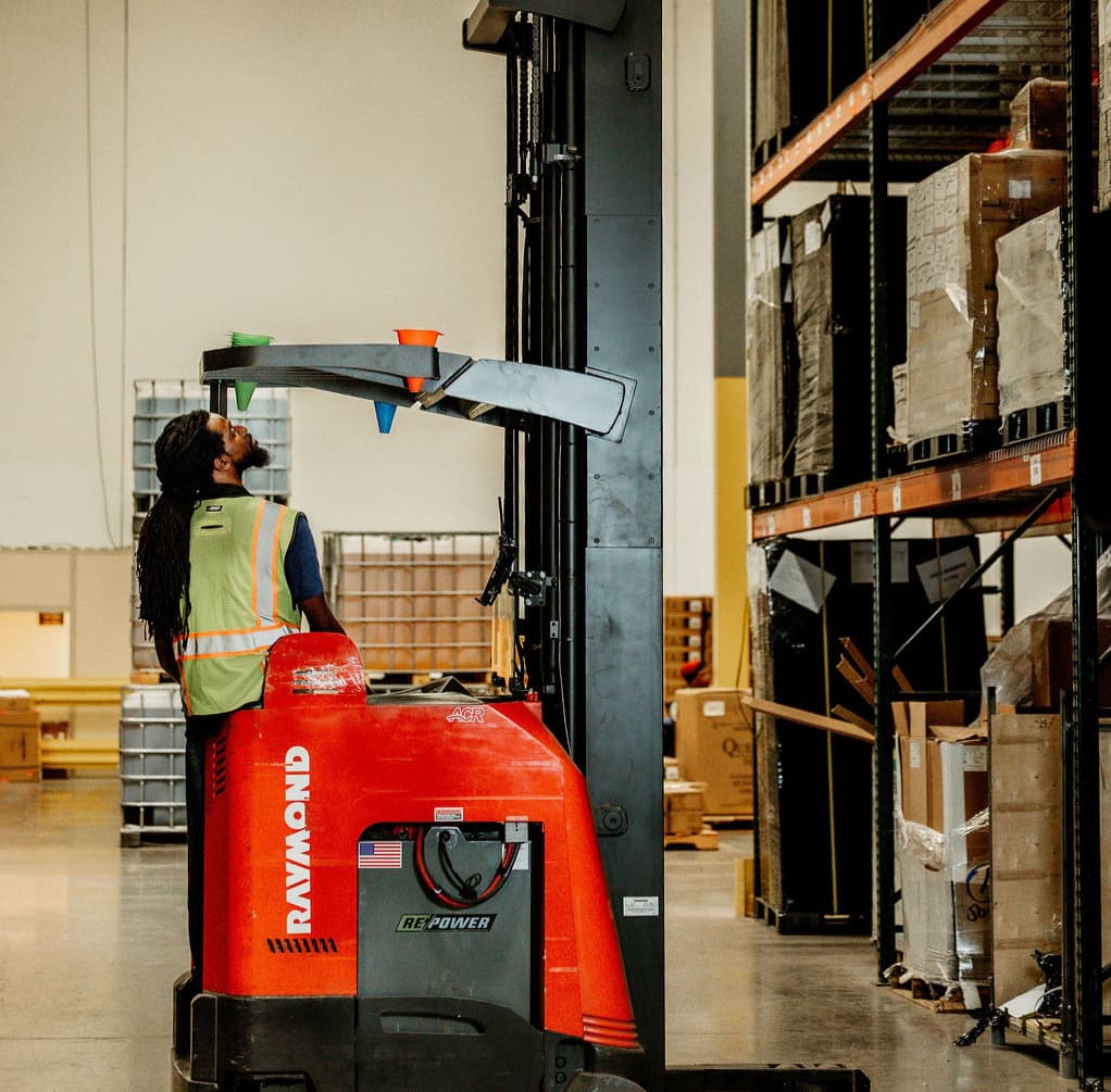A warehouse worker in a reflective safety vest stands beside a red Raymond forklift, looking up at colored loading funnels attached to its mast amid rows of pallet racks