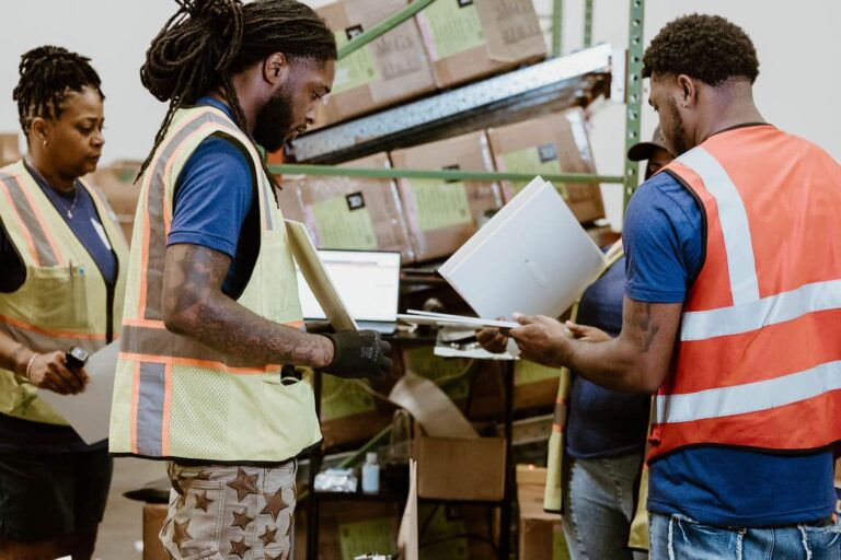 Three warehouse employees in reflective safety vests check inventory on laptops among shelves stacked with cardboard boxes